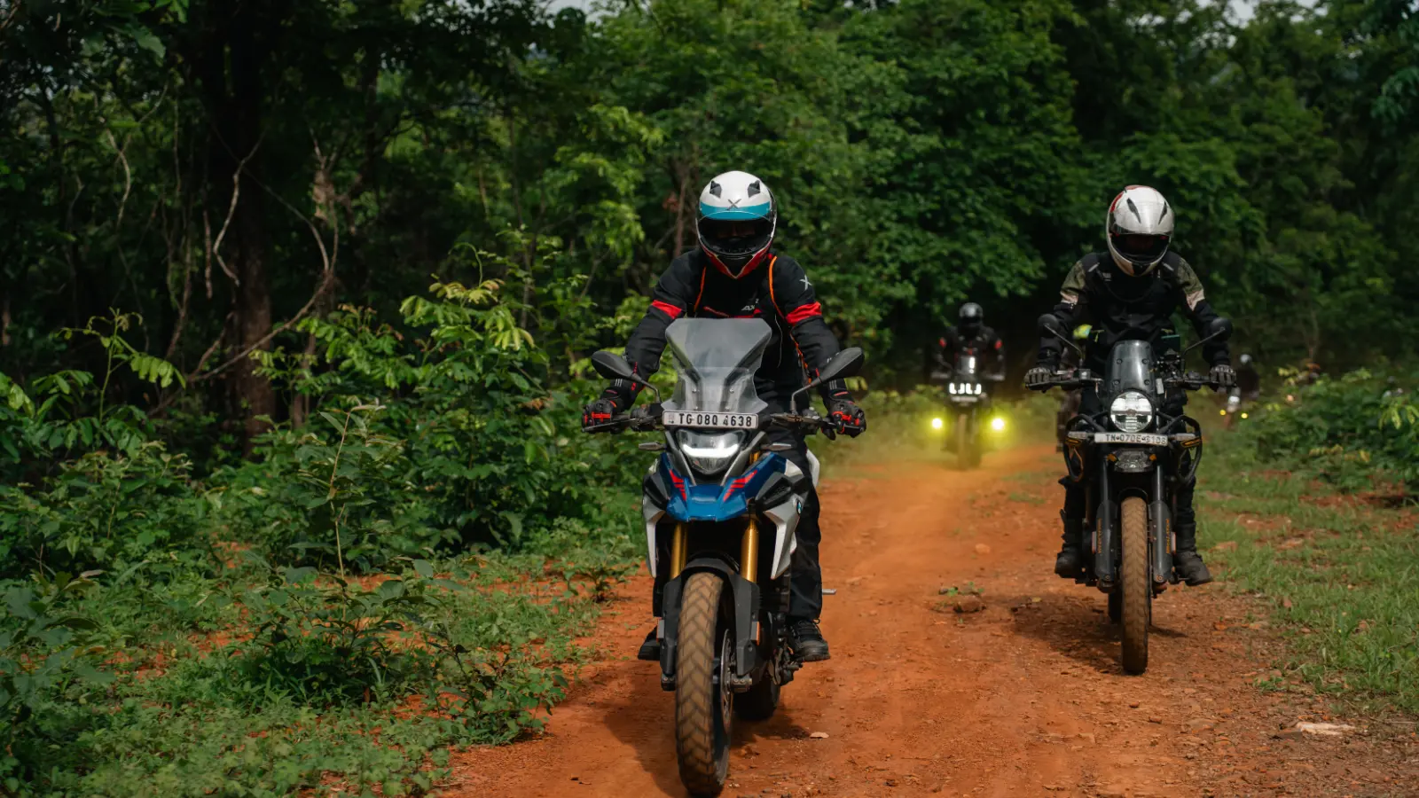 Riders tackle a red dirt forest trail while standing on the foot pegs during a group motorcycle ride.