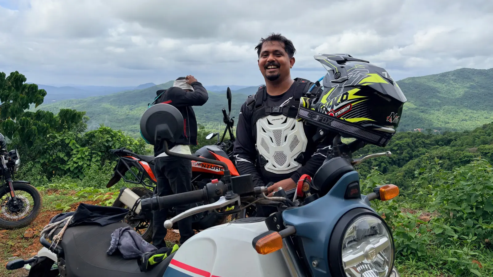 A smiling rider stands beside his motorcycle at a scenic viewpoint after an off road riding session in the Western Ghats.